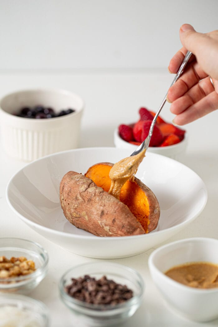 Hand adding a spoonful of peanut butter to a baked sweet potato in a bowl.