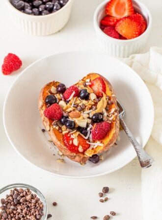 Cut open baked sweet potato in a bowl topped with berries, peanut butter, nuts and coconut.