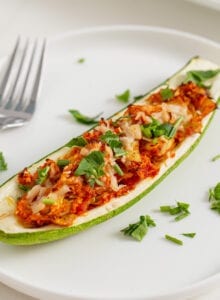 Stuffed zucchini boat topped with cheese and herbs on a plate. Fork rests on plate.
