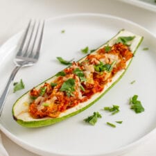 Stuffed zucchini boat topped with cheese and herbs on a plate. Fork rests on plate.