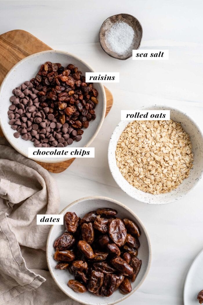 Overhead view of dates, oats, chocolate chips and raisins in bowls. Each bowl is labelled with text overlay.