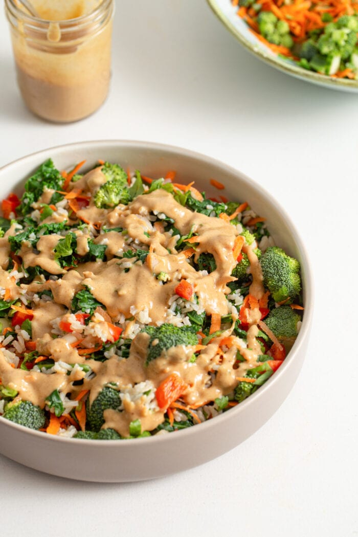 Bowl of kale broccoli brown rice salad topped with peanut sauce.