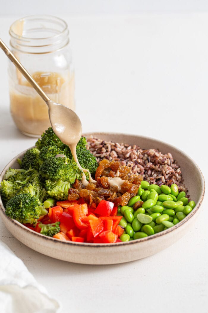 Drizzling a spoonful of tahini dressing over a large bowl of chopped vegetables, edamame and wild rice.
