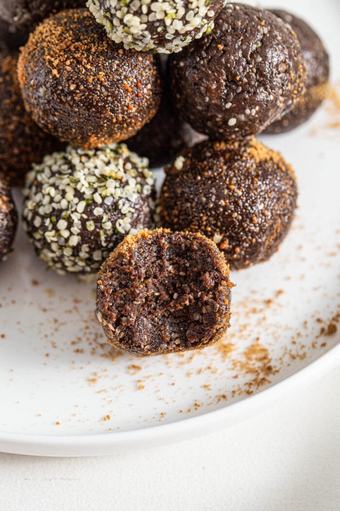 Close up of a superfood chocolate energy ball coated in coconut sugar with a bite taken out of it on a plate. There are more chocolate energy balls on plate in background, some are coated in hemp seeds.