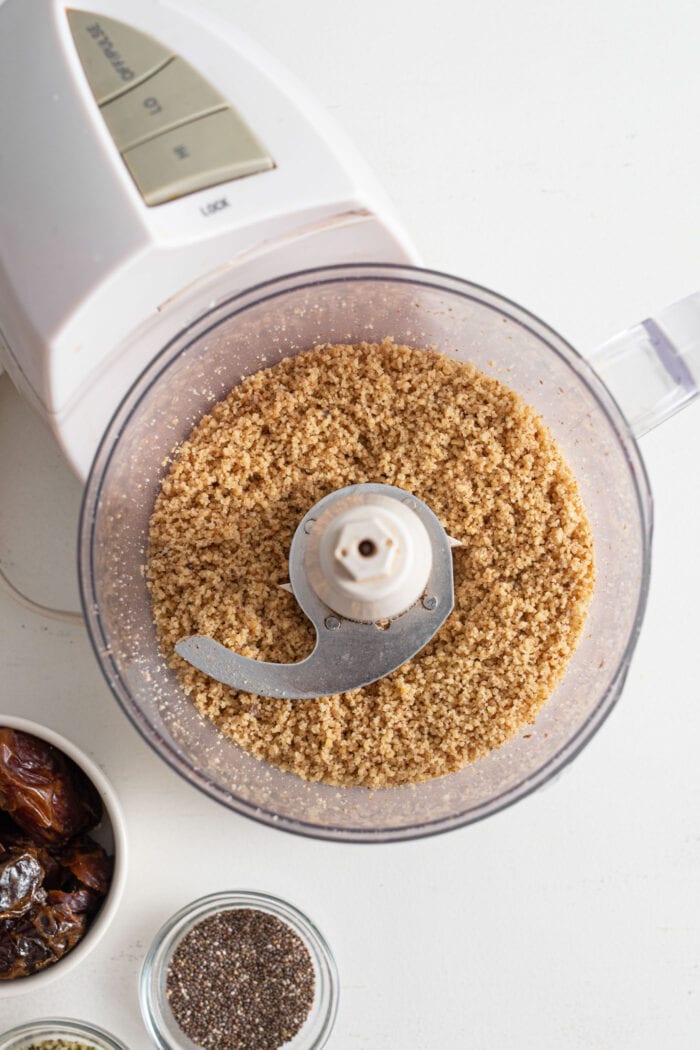Blended walnuts in a food processor. A small glass bowl of chia seeds and dates is beside the food processor.