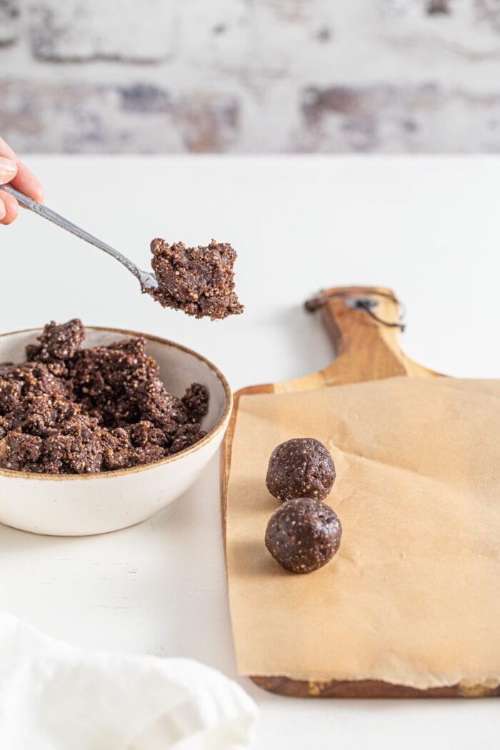 Spoonful of raw chocolate energy ball dough over a cutting board with 2 rolled balls sitting on it.