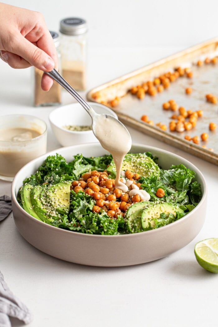 Hand drizzling a spoonful of tahini over a chickpea avocado kale salad.