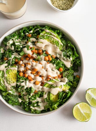 A kale salad with avocado, tahini sauce, chickpea and hemp seeds in a bowl with two pieces of lime beside it.