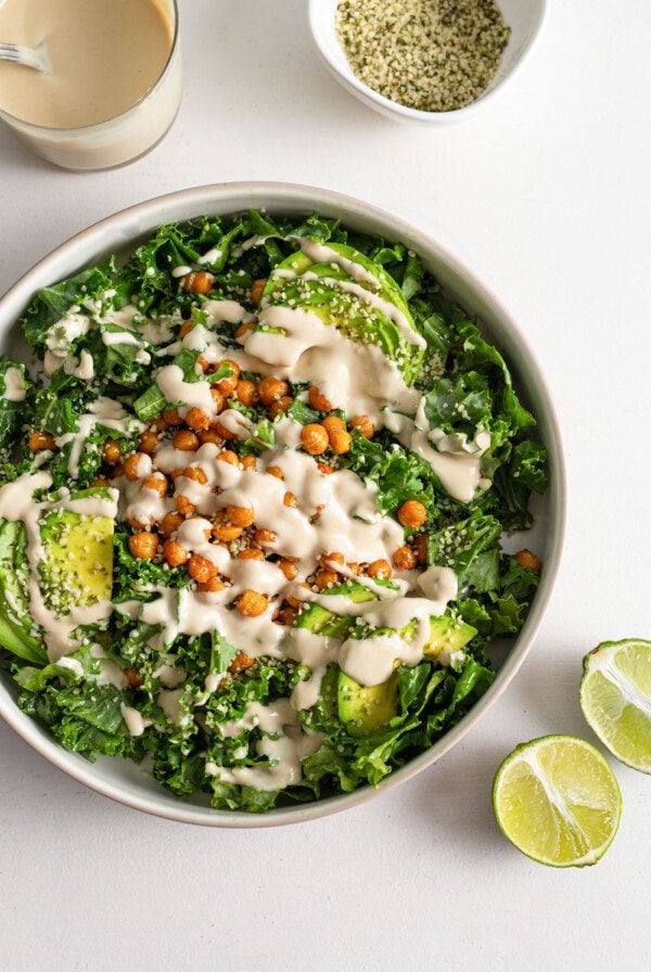 A kale salad with avocado, tahini sauce, chickpea and hemp seeds in a bowl with two pieces of lime beside it.