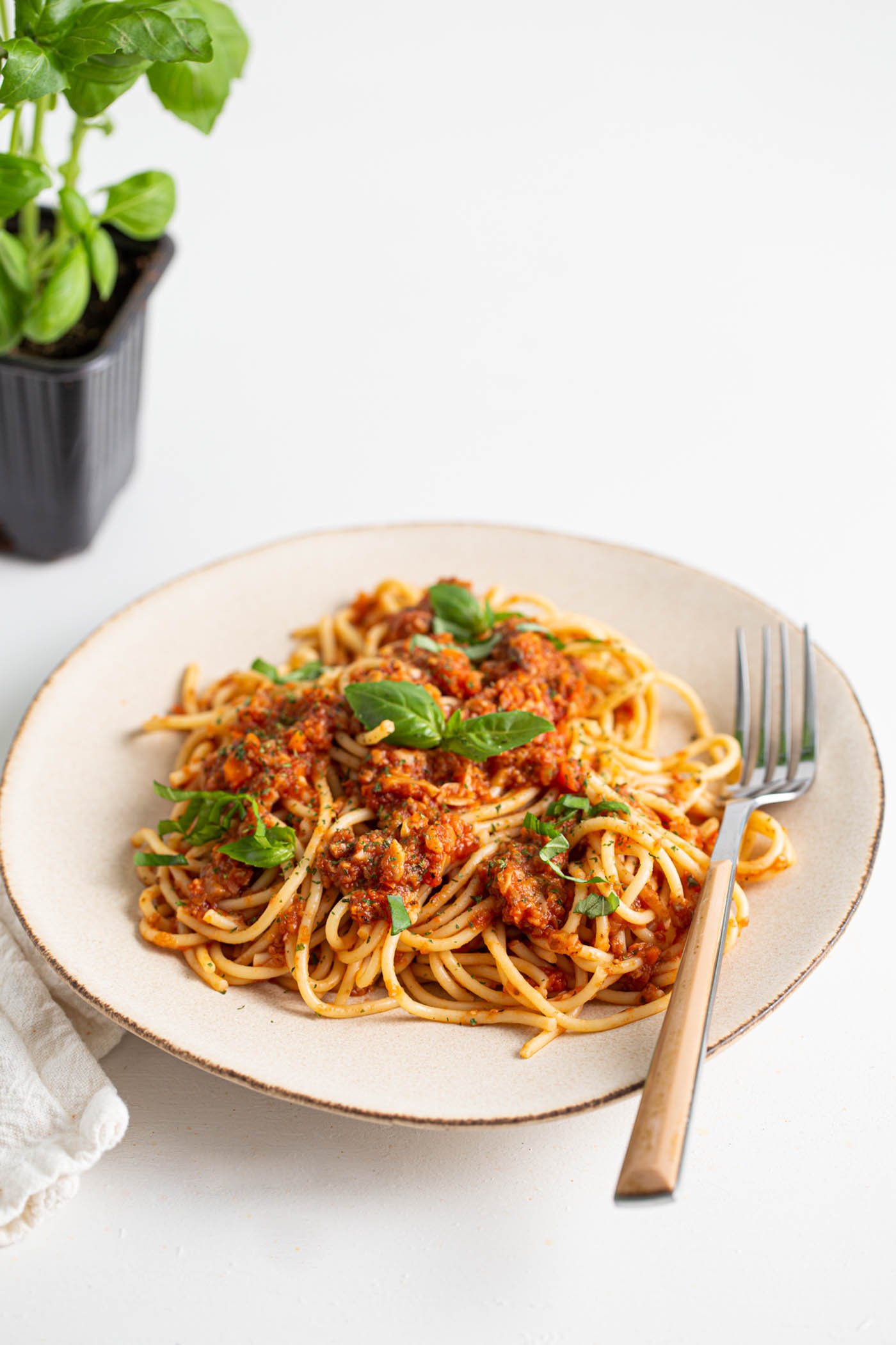 Plate of vegan mushroom bolognese sauce over spaghetti topped with basil and parmesan. A fork rests on the plate and there's a fresh basil plant in the background.