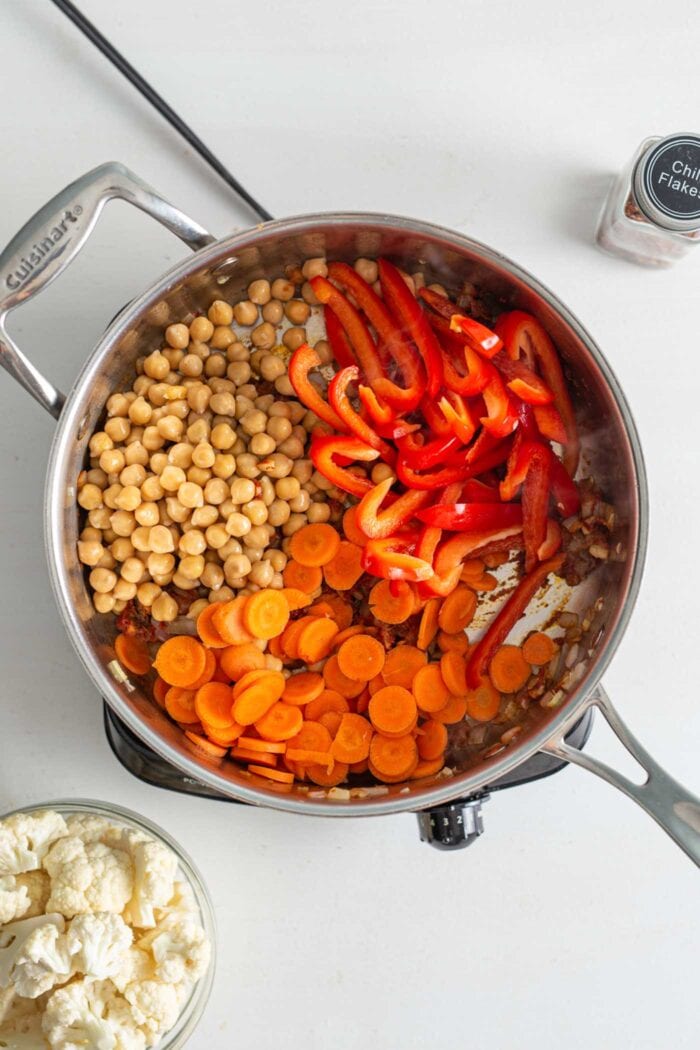 Chickpeas, chopped carrots and sliced bell peppers in a skillet cooking on a small stovetop.