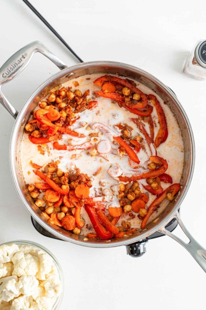 Chickpeas, bell peppers and carrots cooking in coconut milk in a skillet on a small cook top.