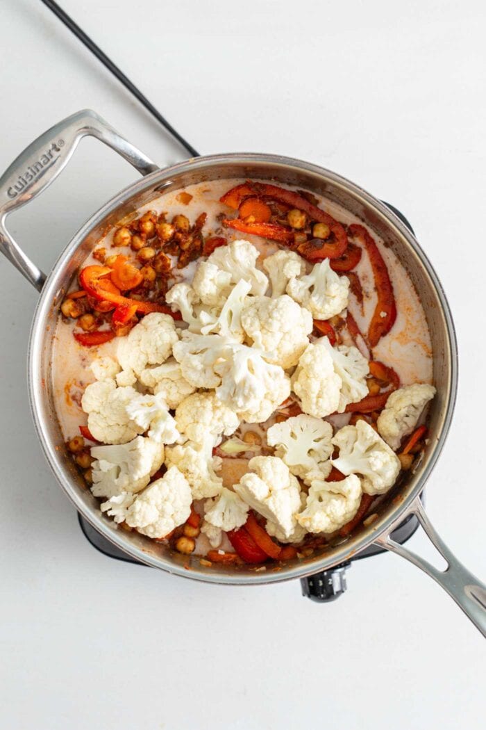 Chickpeas, bell peppers, cauliflower and carrots cooking in coconut milk in a skillet on a small cook top.