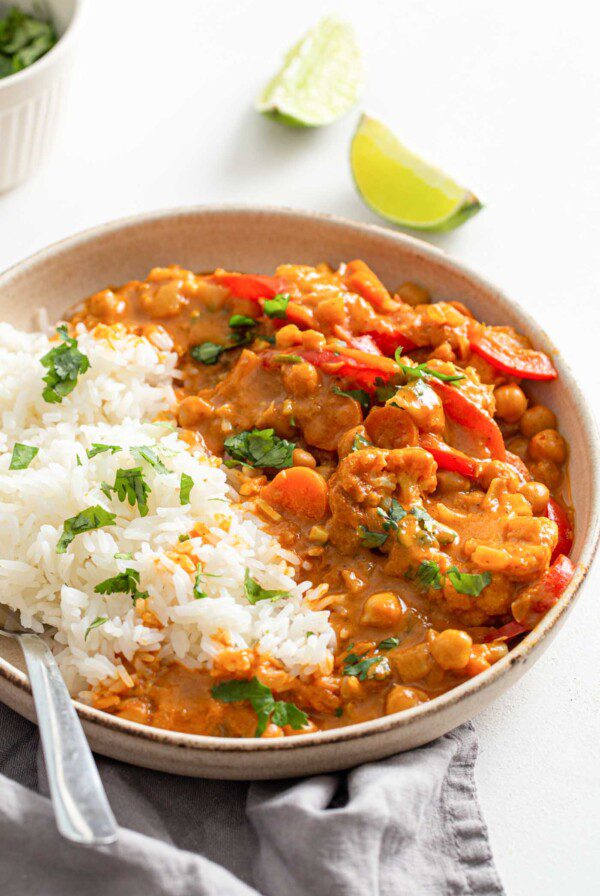 Bowl of rice with cauliflower chickpea curry topped with fresh cilantro. Lime slices beside bowl and small dish of cilantro in background.