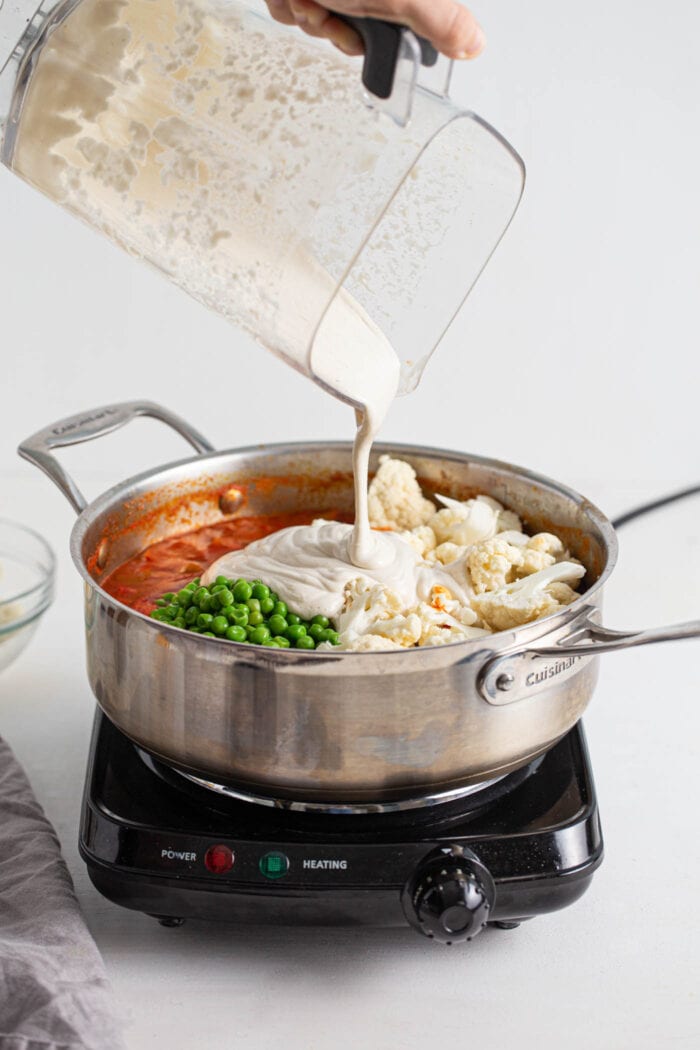 Blender pouring cashew cream into a skillet of tomato, cauliflower and peas.