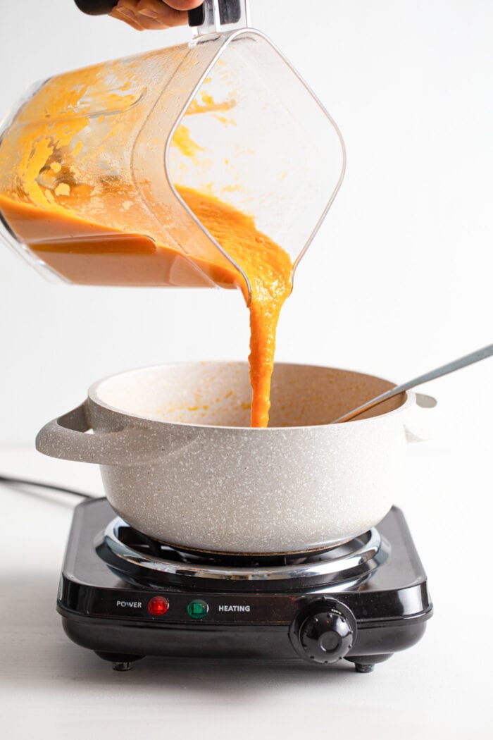 Pouring blended creamy sweet potato corn soup from a blender into a soup pot on a small cooktop.