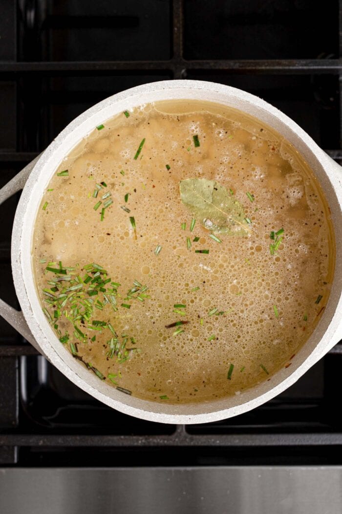 Brothy soup with rosemary cooking in a large pot on a gas range stovetop.