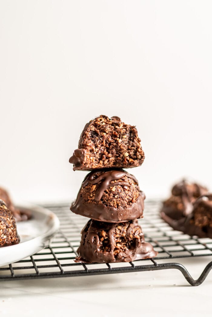 Stack of 3 chocolate dipped chocolate macaroons on a baking cooling rack. The one on top has a bite out of it so you can see the texture inside with little bits of coconut.