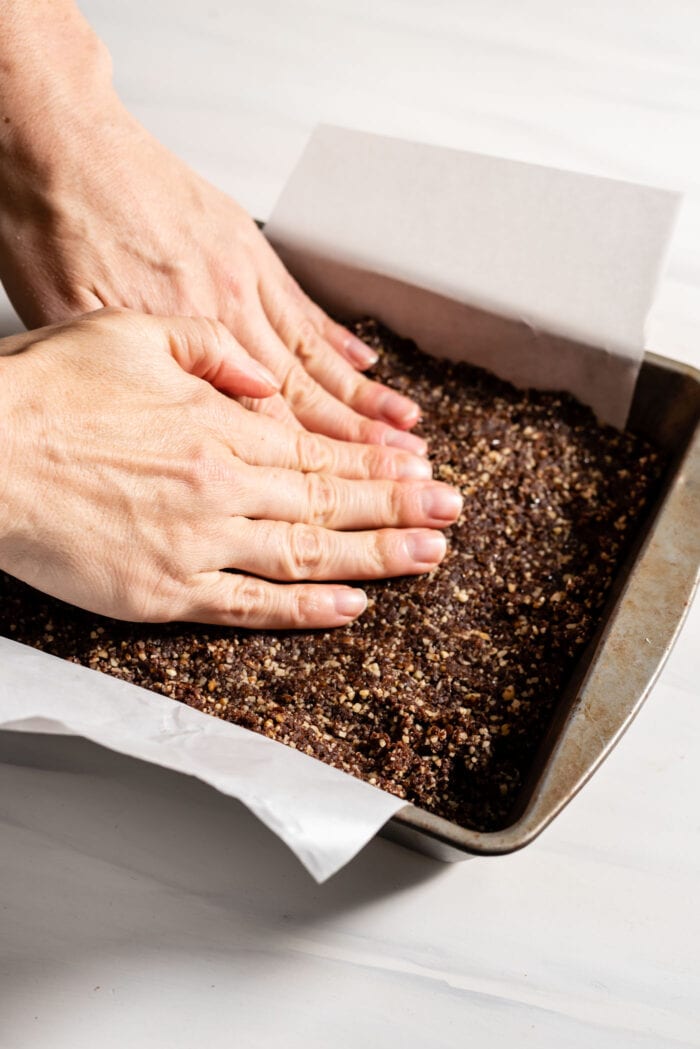 Hands pressing chocolate dough into a small square baking tray lined with parchment paper.