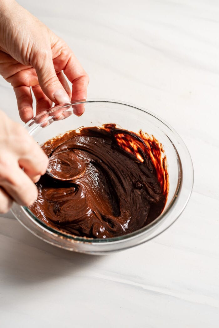 Hands using a spoon to mix a small glass bowl of melted dairy-free chocolate mixed with coconut milk..