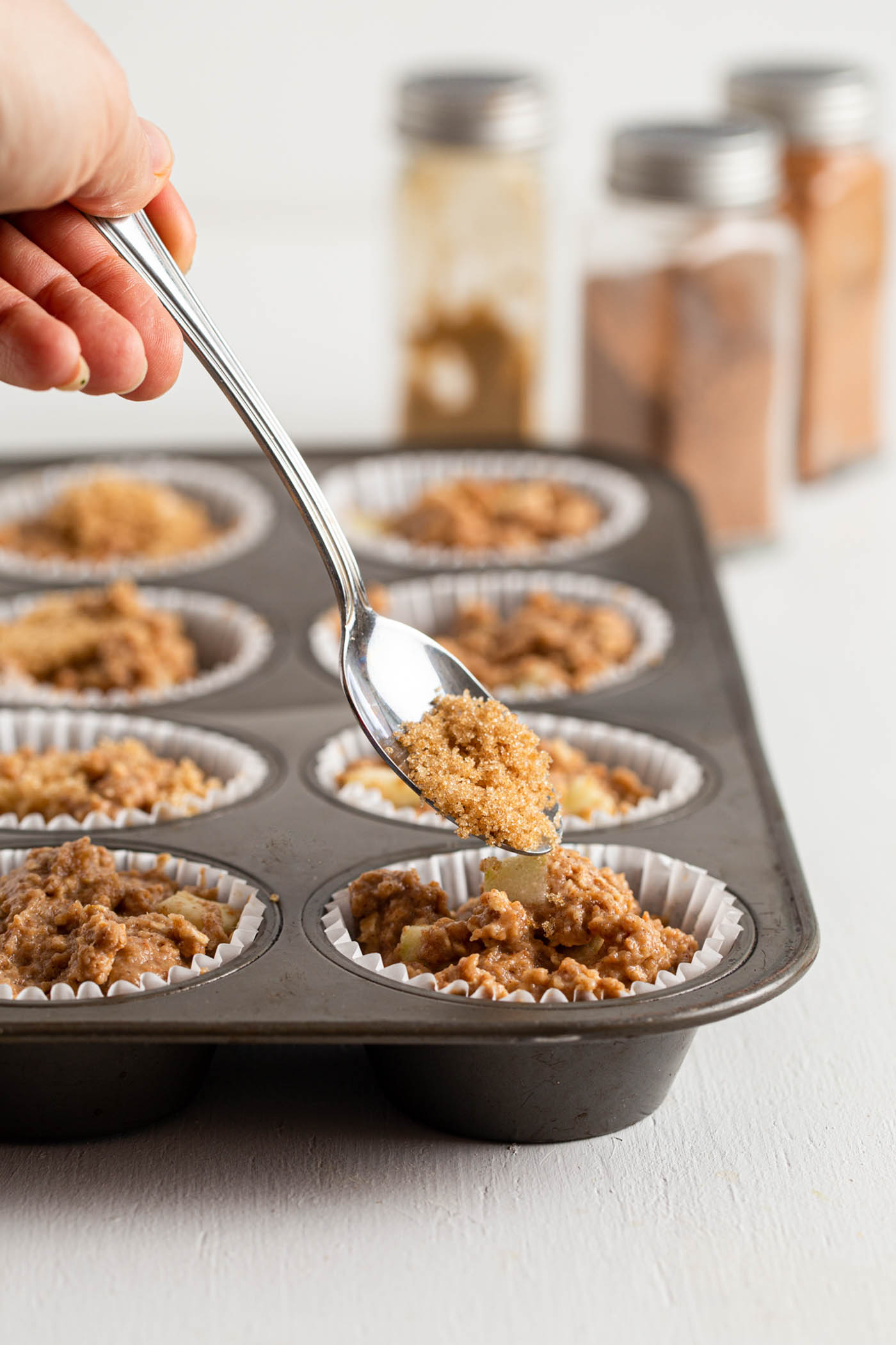 Spooning brown sugar onto a raw muffin in a muffin tin.