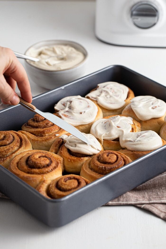 Hand holding a knife and frosting a baking dish of cinnamon rolls.
