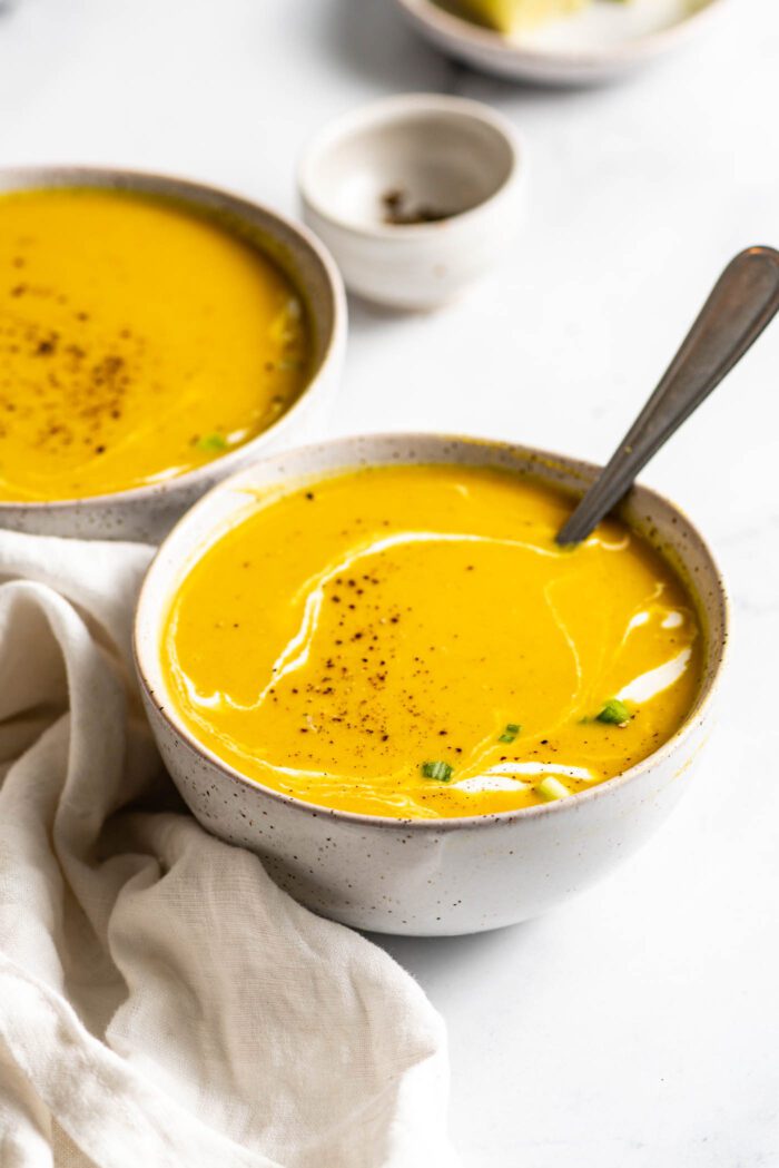 Bowl of squash soup with a drizzle of cream in it. A spoon rests in the bowl and there is another bowl of soup in the background.