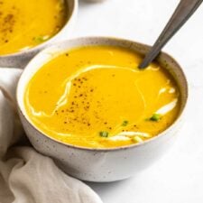 Bowl of squash soup with a drizzle of cream in it. A spoon rests in the bowl and there is another bowl of soup in the background.