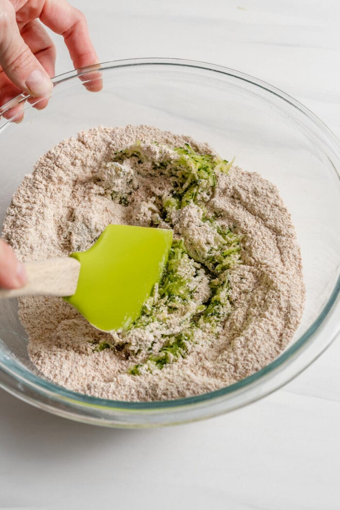 Spatula mixing grated zucchini into a bowl of flour.