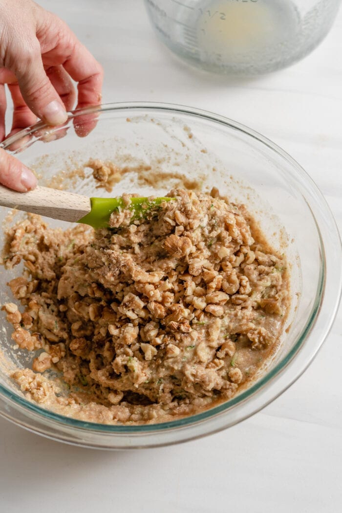 Folding walnuts into raw zucchini bread batter in a large mixing bowl.