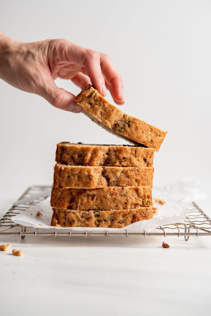 Hand picking up a slice of zucchini loaf from a stack of slices sitting on a cooling rack.