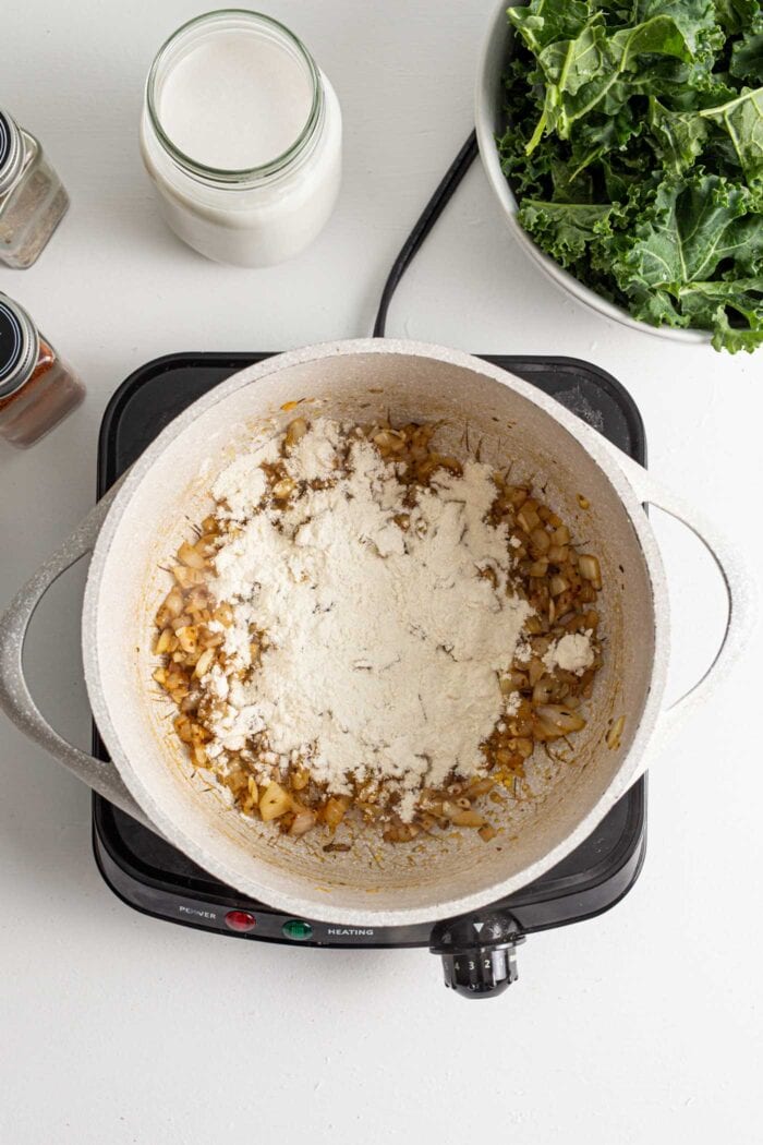 Flour sprinkled on onions in a pot on a hot plate with a bowl of kale beside it..