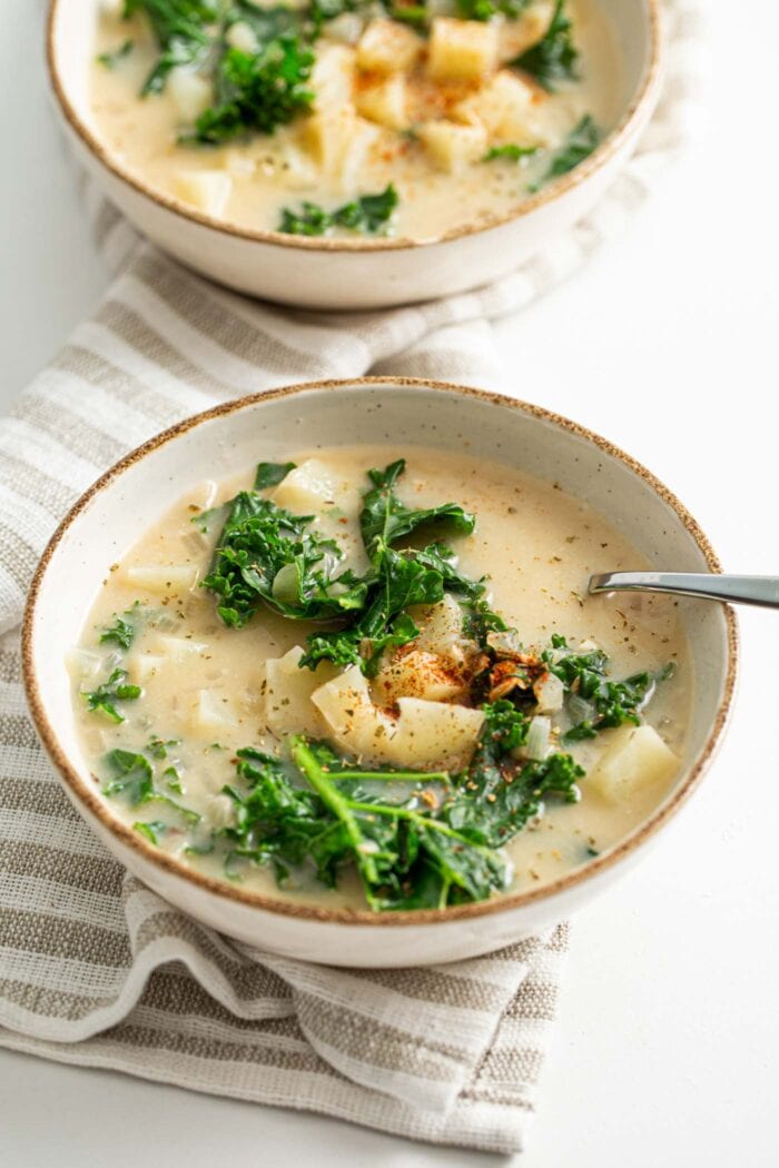 Two bowls of creamy vegan zuppa Toscana soup with chunks of potato and kale. A spoon rests in one of the bowls.