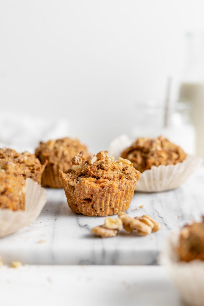 A carrot muffin on a marble cutting board with more muffins in the background and some walnuts scattered around.
