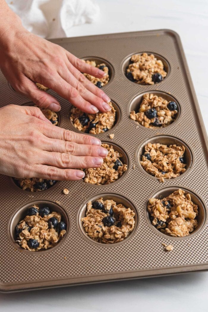 Two hands pressing oatmeal cup dough into a 12-cup muffin tin.