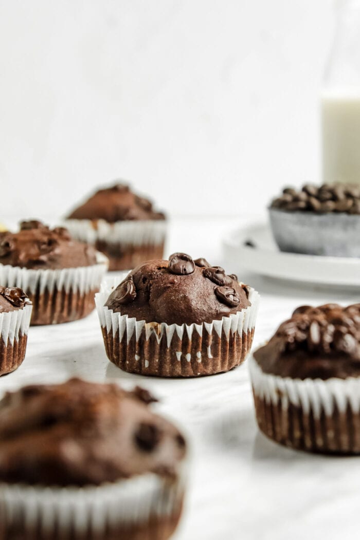 A double chocolate muffin in a cupcake liner sitting on a marble surface with a number of other muffins around it out of focus.