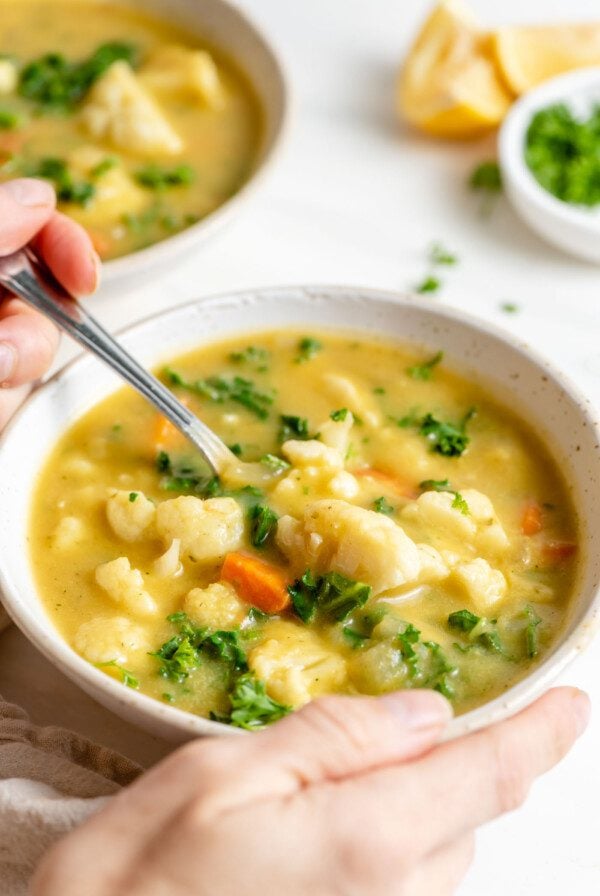 Hand using a spoon to take a spoonful of cauliflower soup from a bowl.