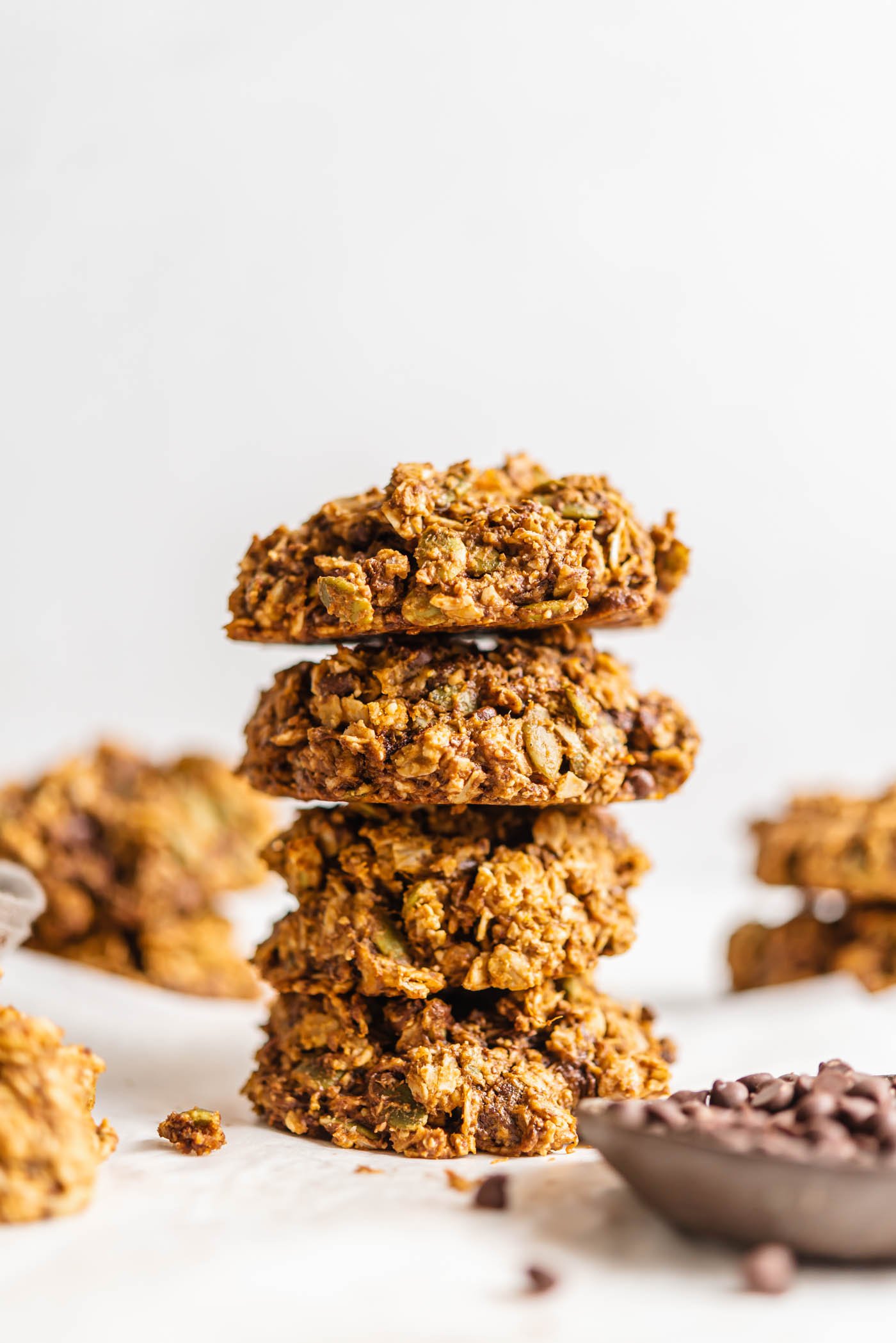 Stack of 4 hearty cookies with a small dish of chocolate chips in foreground and more cookies in background.