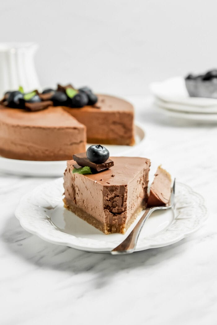 Slice of chocolate cheesecake with a fork resting beside it on a small decorate plate and a whole cheesecake in the backgorund.