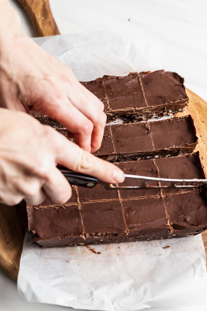 Hand using a knife to cut chocolate fudge bars into 16 squares on a cutting board.
