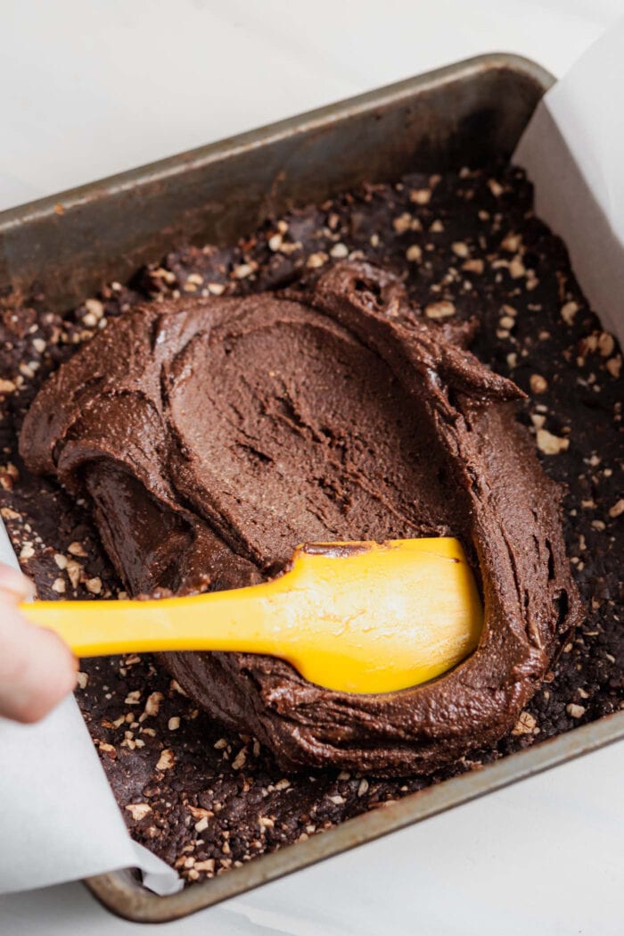 Using a small spatula to spread thick chocolate fudge over a crust layer in a square baking pan.
