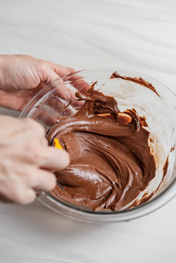 Hands using a spatula to mix melted chocolate in a glass mixing bowl.