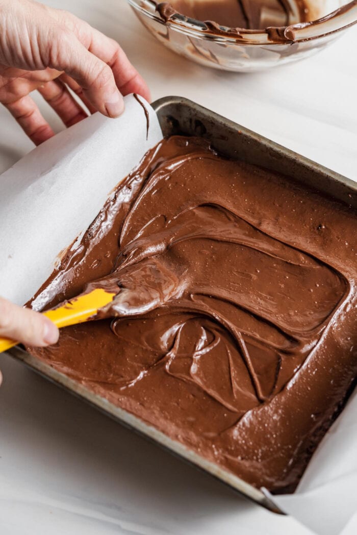 Hand using a spatula to spread melted chocolate over a bars in a square baking pan.
