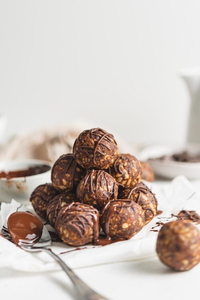Stack of no-bake energy balls drizzled in chocolate sitting on a folded piece of parchment paper beside a spoon with melted chocolate on it.