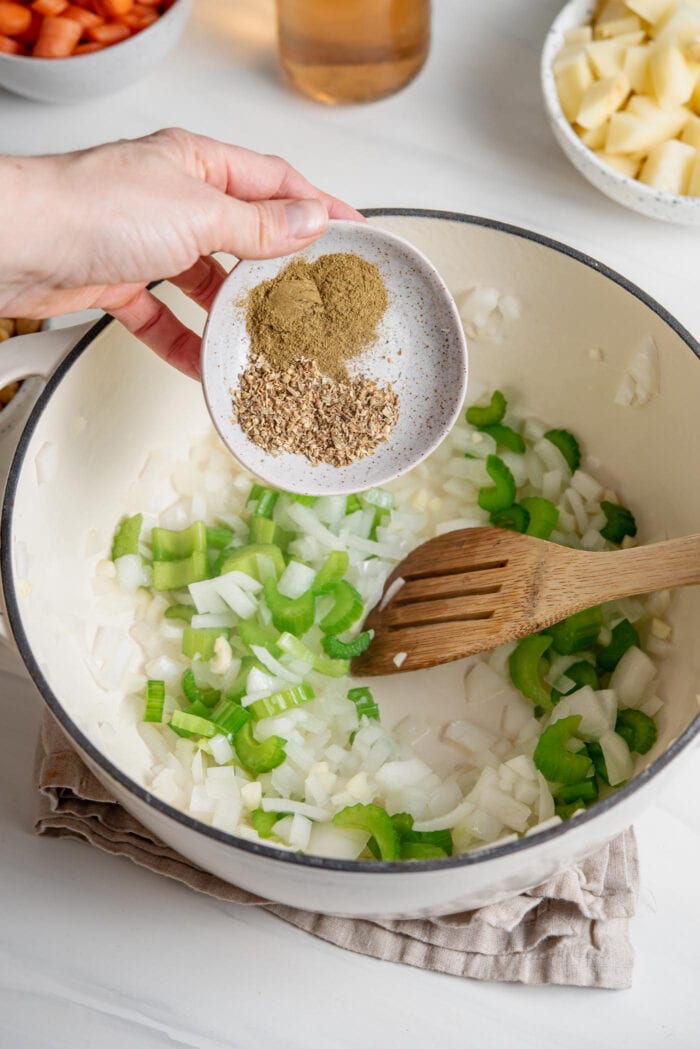Adding a small dish of dried herbs to celery and onion cooking in a pot.
