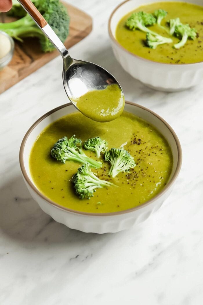 A spoon dipping into a small bowl of cream of broccoli soup topped with broccoli pieces and black pepper.