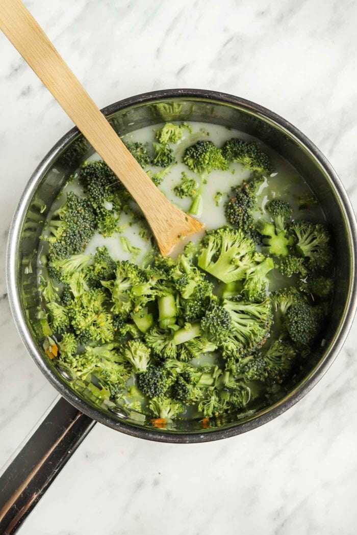 Chopped broccoli cooking in milk in a wooden pot with a spoon in it.