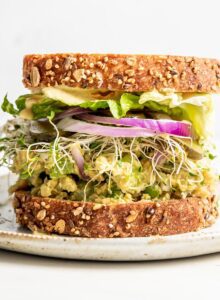 Front view of a chickpea salad sandwich with sprouts, pickles and red onion on it.