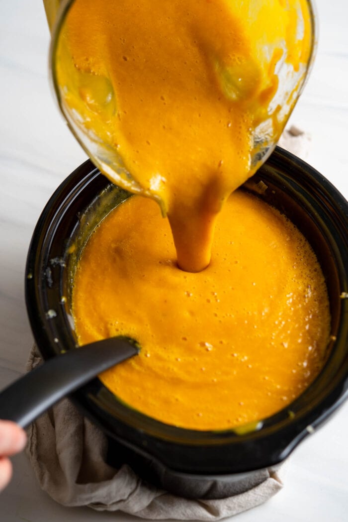 Pouring creamy butternut squash soup from a blender into a slow cooker.