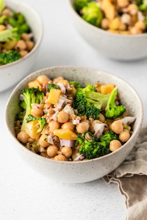 Bowl of chickpea salad with broccoli, farro, bell peppers and red onion. Two more bowls in background.